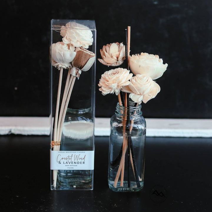 A clear bottle from Cedar Mountain Candle’s SPRING &amp; SUMMER: Flower Reed Diffusers, featuring natural rattan reeds and cream-colored sola wood flower diffusers in the Cedarwood &amp; Lavender scent, sits beside its packaging against a dark background.