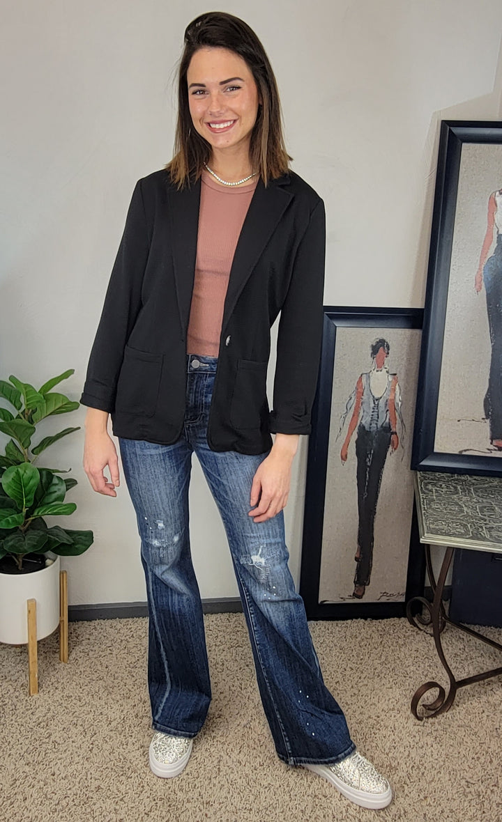 A woman smiles indoors wearing the Doe & Rae Kallie Blazer over a dusty pink top with distressed flare jeans and sparkly sneakers. Framed fashion sketches and a potted plant provide the backdrop.