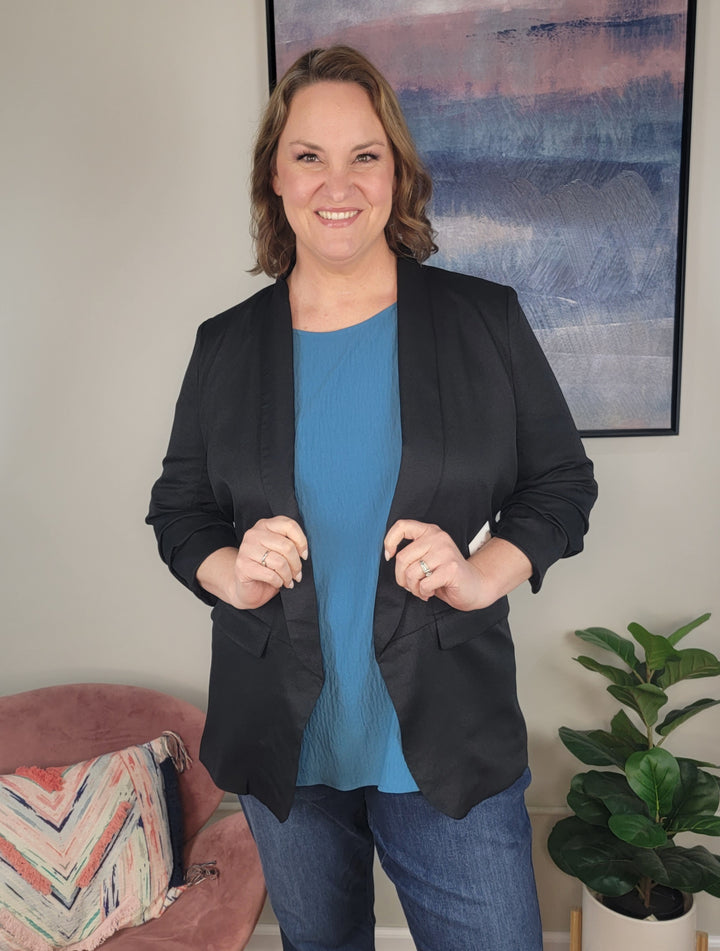 A smiling woman with shoulder-length brown hair stands indoors, wearing the fully lined Zoie Blazer by ODDI—featuring ruched sleeves—over a blue top and jeans. Behind her are a framed abstract painting, a pink chair with a patterned pillow, and a potted plant.