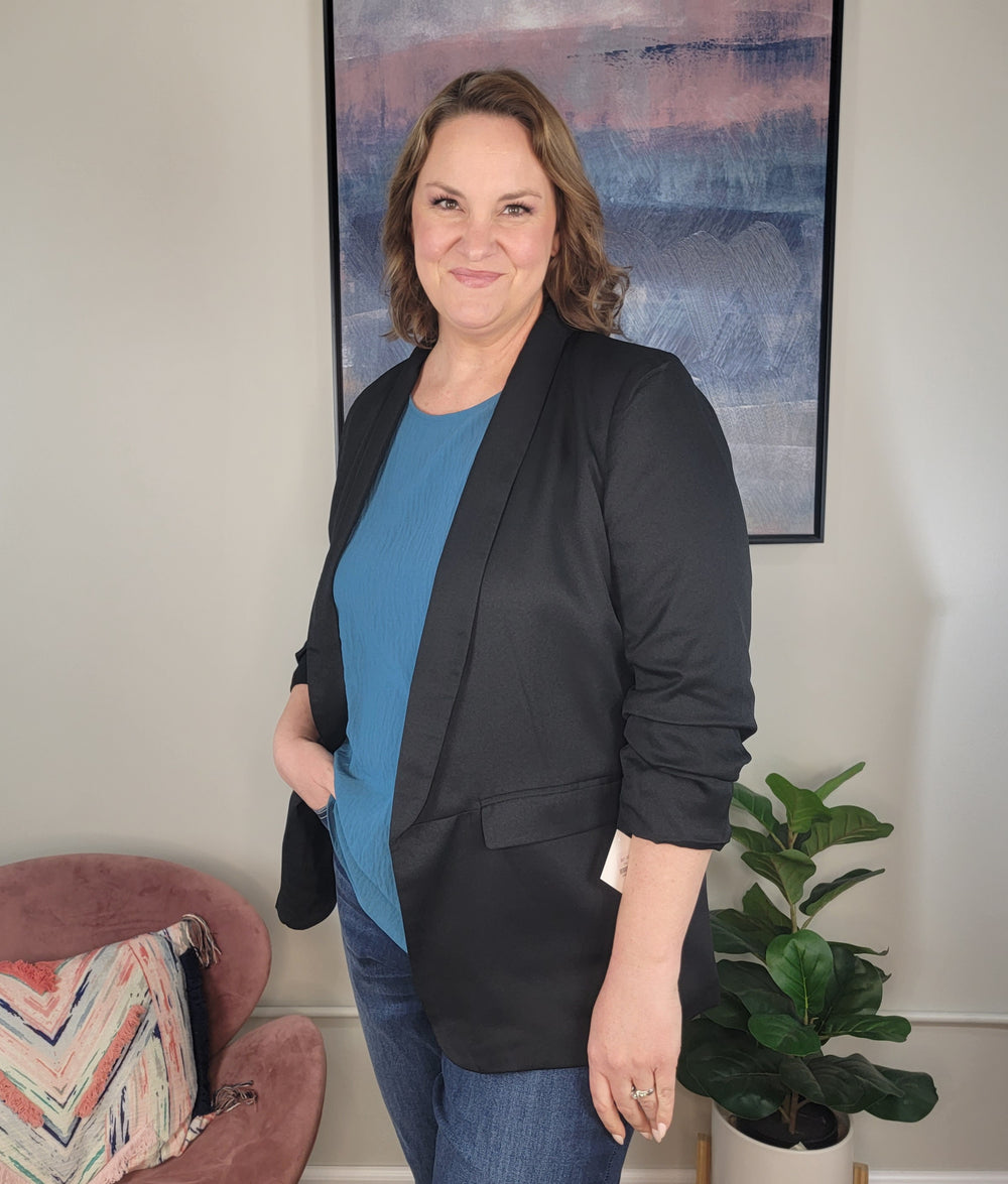 A woman with shoulder-length brown hair smiles indoors, wearing the ODDI Zoie Blazer over a blue top and jeans. In the background are a plant, a pink chair with a patterned pillow, and abstract wall art.