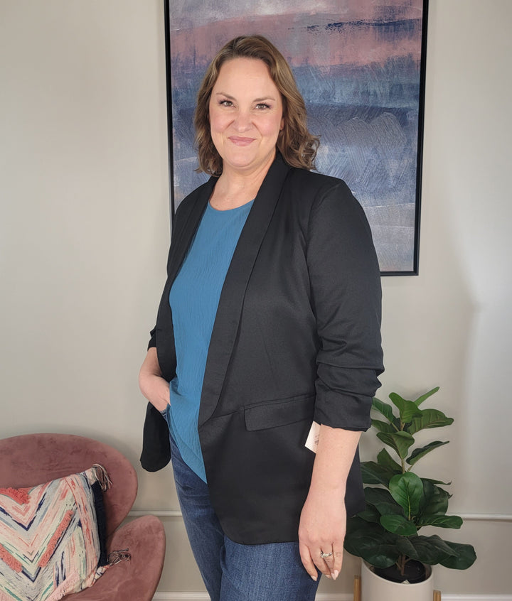 A woman with shoulder-length brown hair smiles indoors, wearing the ODDI Zoie Blazer over a blue top and jeans. In the background are a plant, a pink chair with a patterned pillow, and abstract wall art.