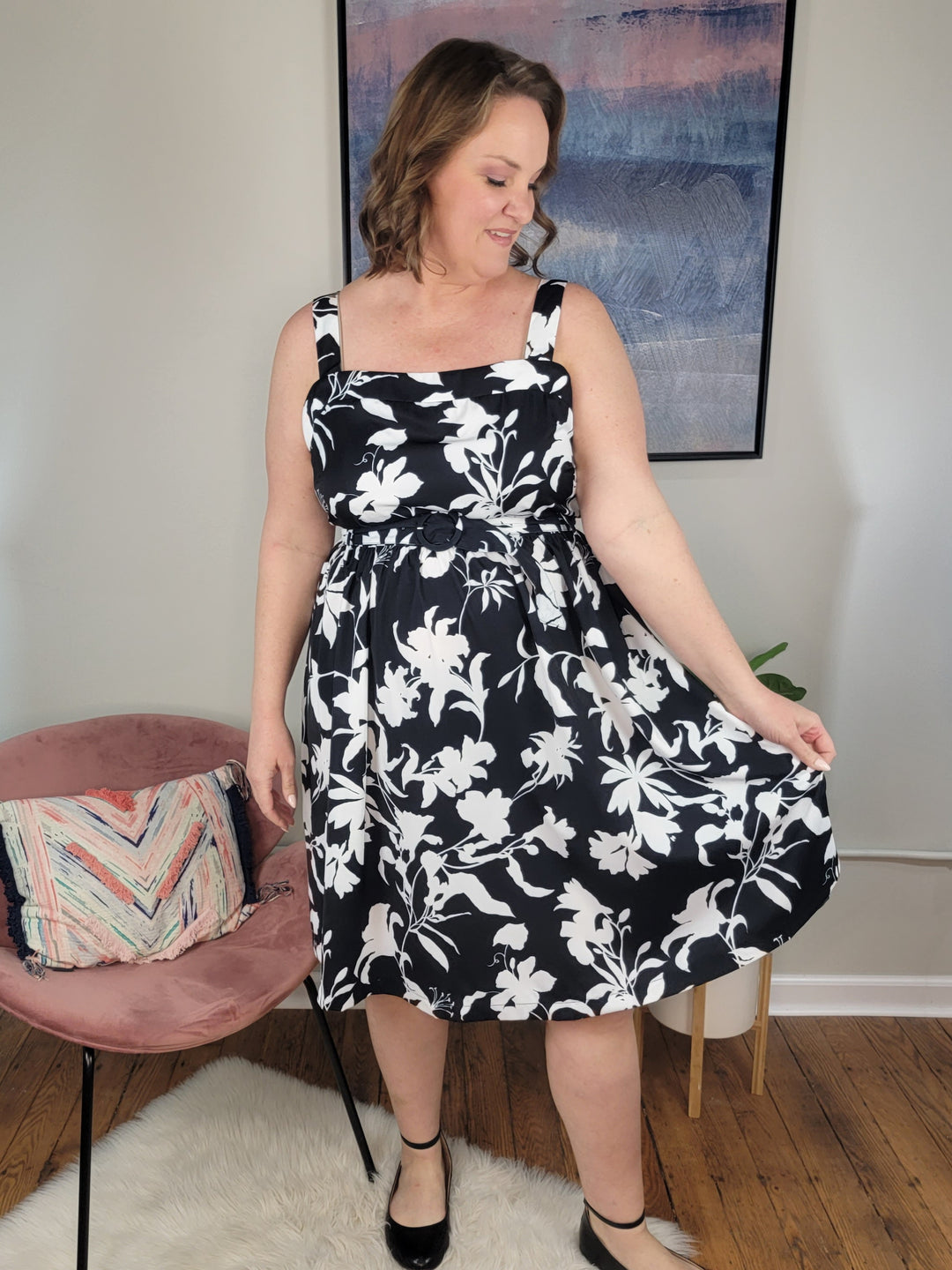 A woman wears the ODDI Angela Dress, featuring a belted waist and floral print, as she stands indoors smiling and holding her skirt. Behind her are a pink chair with a decorative pillow and framed abstract art on the wall.