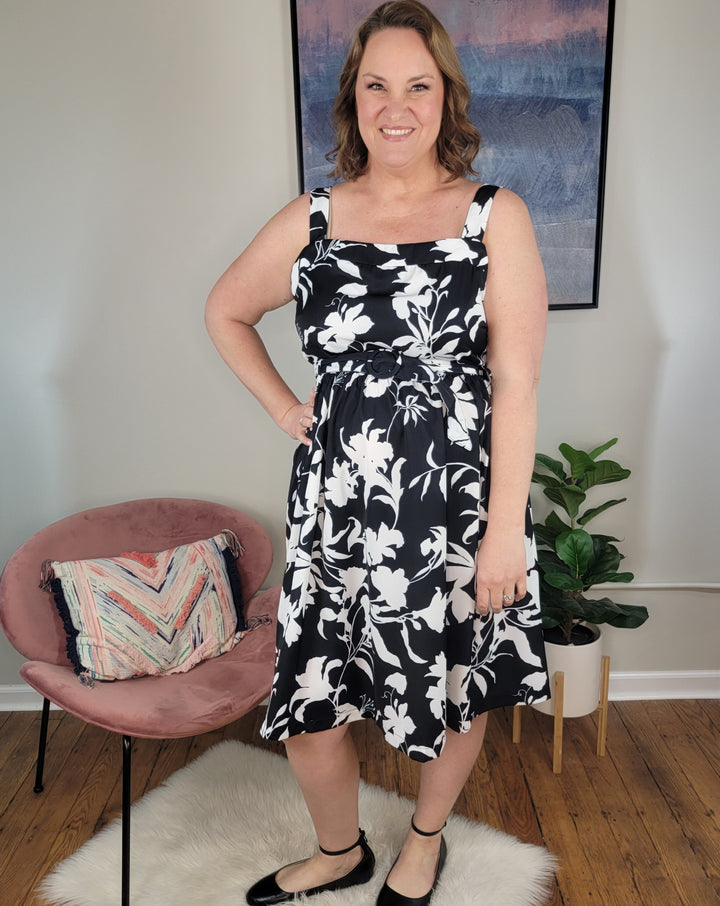 A woman stands indoors on a white rug, smiling and wearing the ODDI Angela Dress with black shoes. Behind her are a pink chair with a colorful pillow, a green potted plant, and a framed picture on the wall.