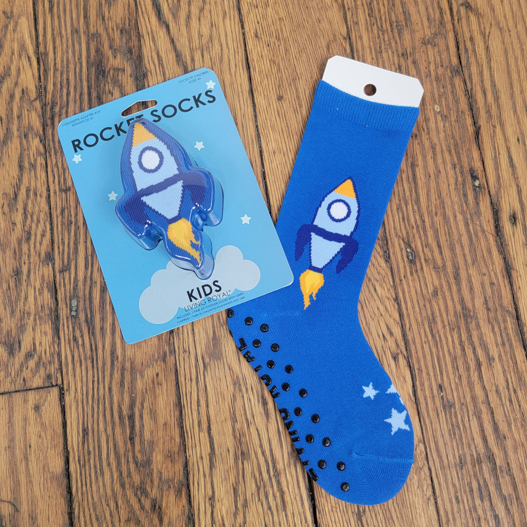 Living Royal Kids: 3D Socks feature a blue, non-slip design with white stars and a fun rocket pattern; one sock comes in rocket-shaped packaging, while the other is displayed flat on a wooden floor for added detail.
