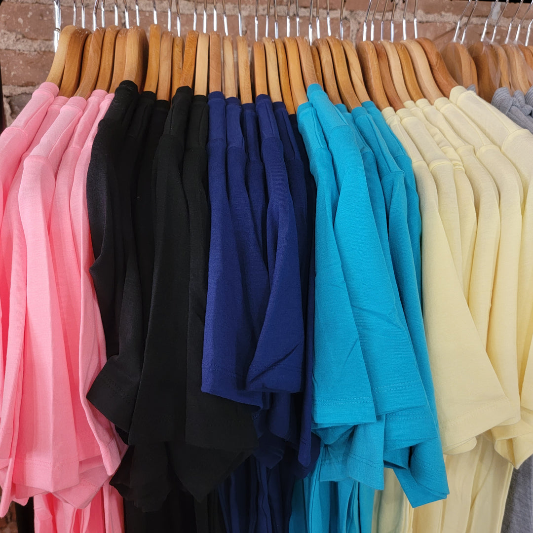 A row of Zenana Basic Tee shirts in pink, black, blue, teal, yellow, and light gray hangs neatly on wooden hangers against a brick wall—ideal as a classic layering top.