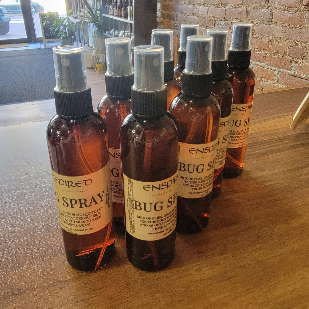 Eight brown Enspired Bug Spray bottles with spray tops are arranged on a wooden table in a store, with brick walls and shelves in the background.