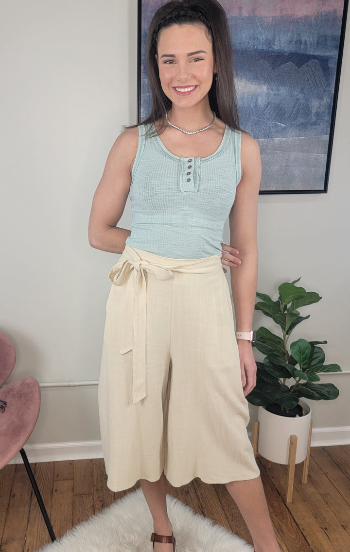A woman smiles indoors, wearing a light blue sleeveless top and LLove Inc Calista Wide Leg Cropped Capris. She stands on a white rug next to a potted plant and pink chair, with artwork displayed on the wall behind her.