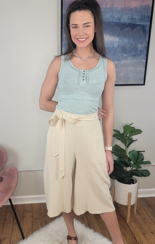 A woman smiles indoors, wearing a light blue sleeveless top and LLove Inc Calista Wide Leg Cropped Capris. She stands on a white rug next to a potted plant and pink chair, with artwork displayed on the wall behind her.