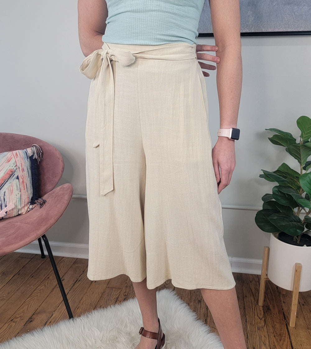A person wearing LLove Inc Calista Wide Leg Cropped Capris, a light blue sleeveless top, brown sandals, and a smartwatch stands on a white rug next to a pink chair and potted plant.