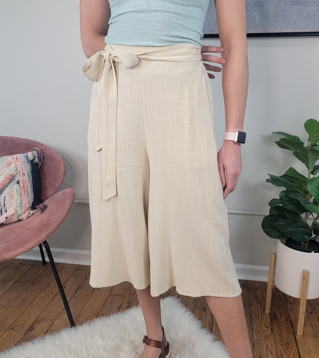 A person wearing LLove Inc Calista Wide Leg Cropped Capris, a light blue sleeveless top, brown sandals, and a smartwatch stands on a white rug next to a pink chair and potted plant.