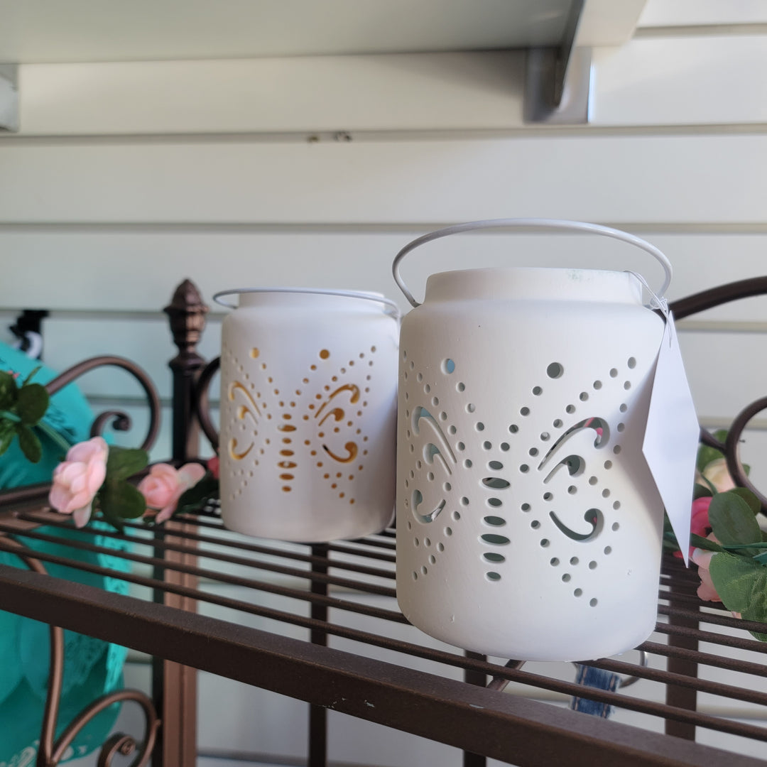 Wills Butterfly Lanterns with delicate cutouts rest on a decorative metal shelf, surrounded by pink artificial flowers and green leaves, creating a beautiful contrast against the white paneled wall.