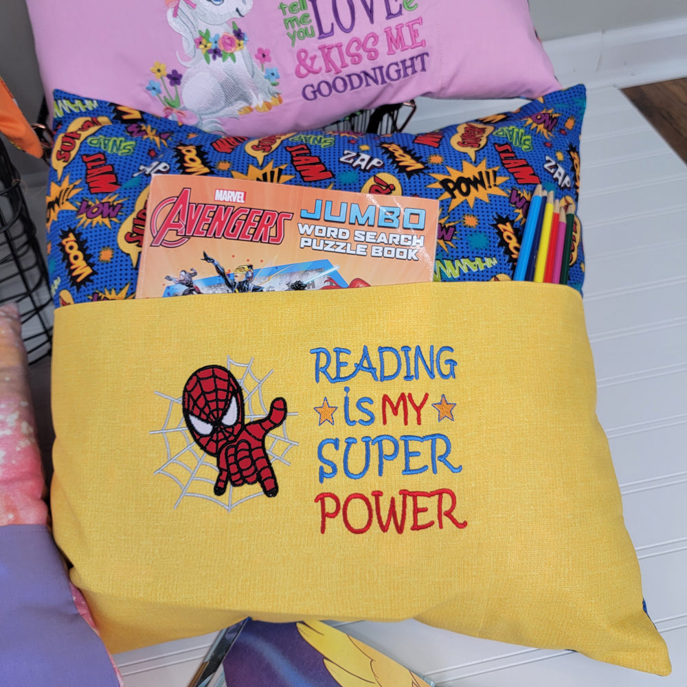The Diana Kuehl Reading Pillow is a handmade yellow pillow for kids, embroidered with Spider-Man, a web, and "Reading is my super power." The pocket stores an Avengers word search book and colored pencils. Other colorful Reading Pillows are visible behind.