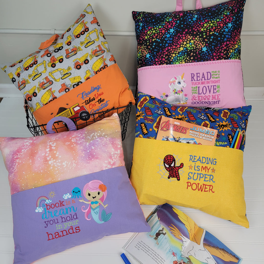 Diana Kuehl Reading Pillows: Four colorful handmade pillows for kids, each with embroidered reading messages and pockets for books or pencils. Designs feature trucks, unicorns, superheroes, mermaids, and stars. Displayed with an open book on a white surface.