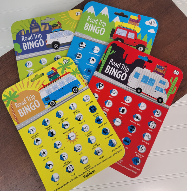 Four colorful Toysmith Road Trip Travel BINGO cards are displayed on a wooden surface. Each game board features fun icons—cars, road signs, animals—and a cheerful cartoon vehicle graphic on top.