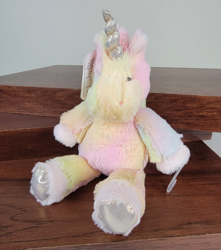 The Mary Meyer Kids Plush Animals unicorn, featuring pastel colors, sparkly silver hooves, and a soft, fluffy texture, sits on wooden steps. With an ear tag and adorable design, it makes a magical gift for children.
