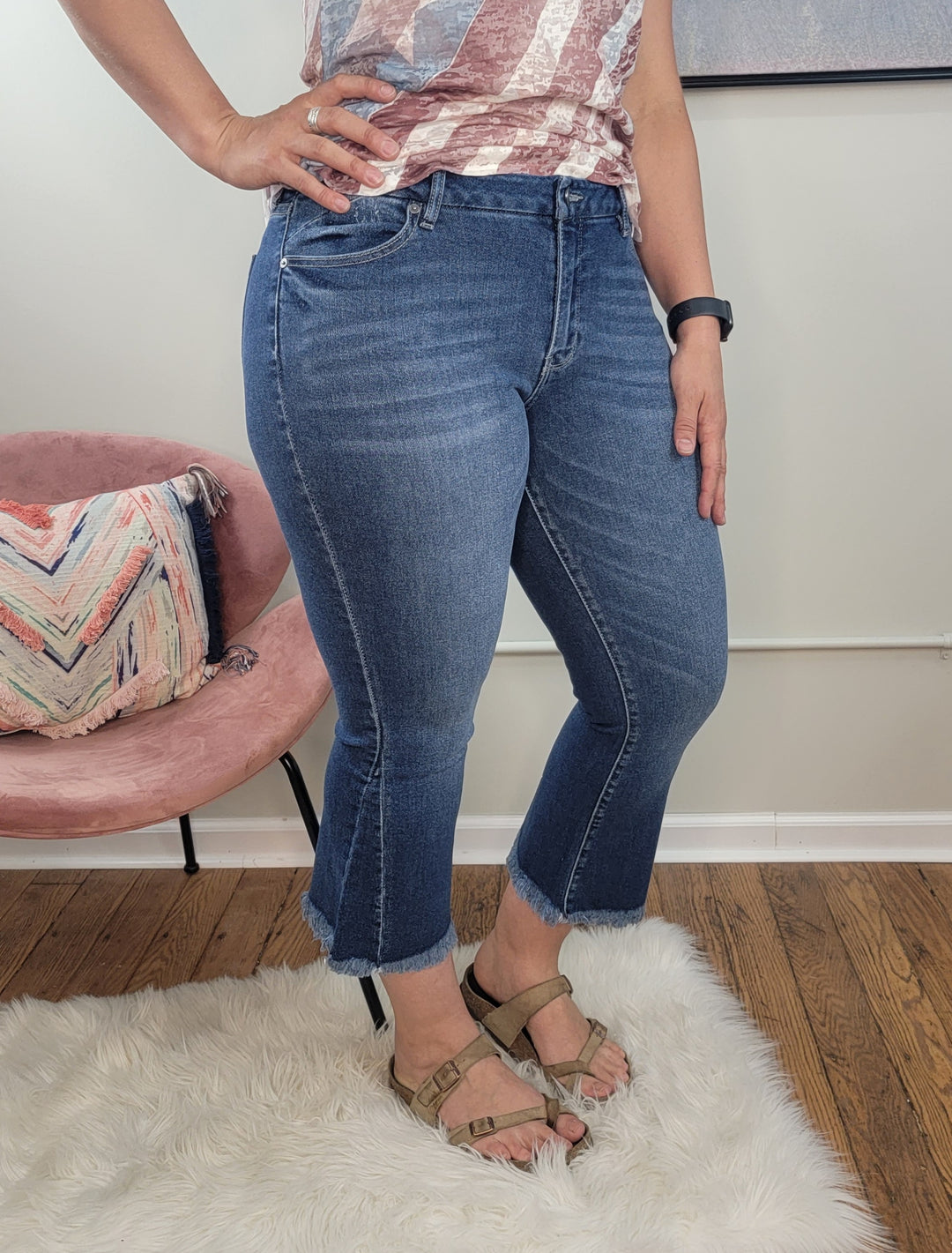 The lower half of a person is shown wearing KanCan's Merlie Cropped Flare jeans with frayed hems and sandals, standing on a white rug in a room featuring a pink chair and chevron-patterned pillow.