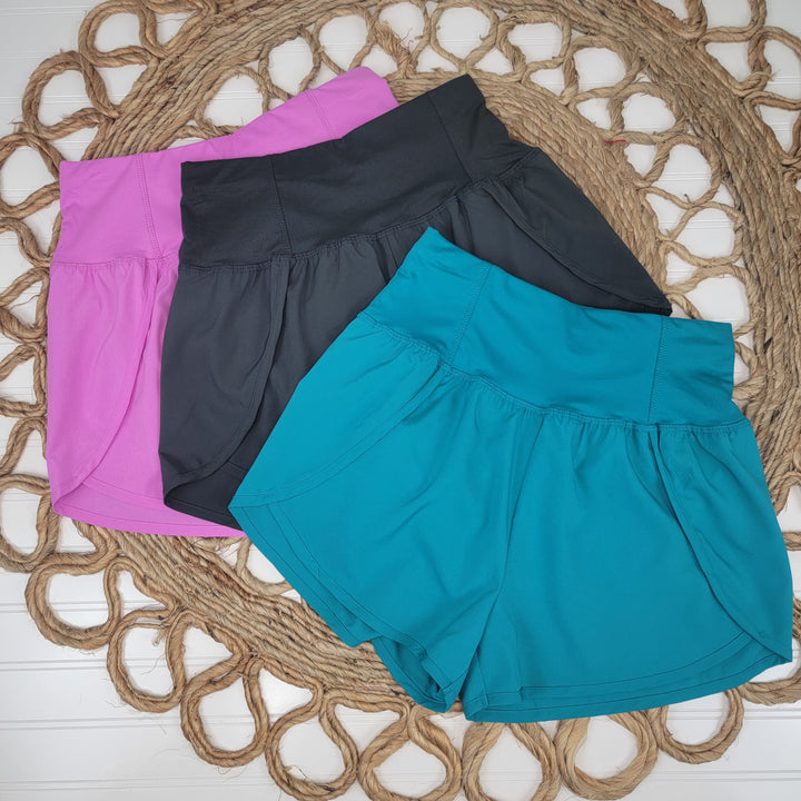 Three pairs of Zenana Adrianne Shorts—high waisted with side slits in purple, black, and teal—are neatly arranged on a round woven rug set against a white wooden background.