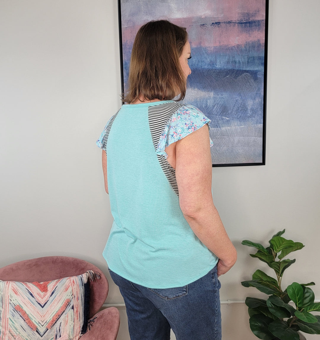A woman faces away from the camera in a modern room, wearing the Emerald Riley Top—a light blue ruffle-sleeve blouse—with blue jeans. A pink chair, vibrant green plant, and abstract painting complete the setting.