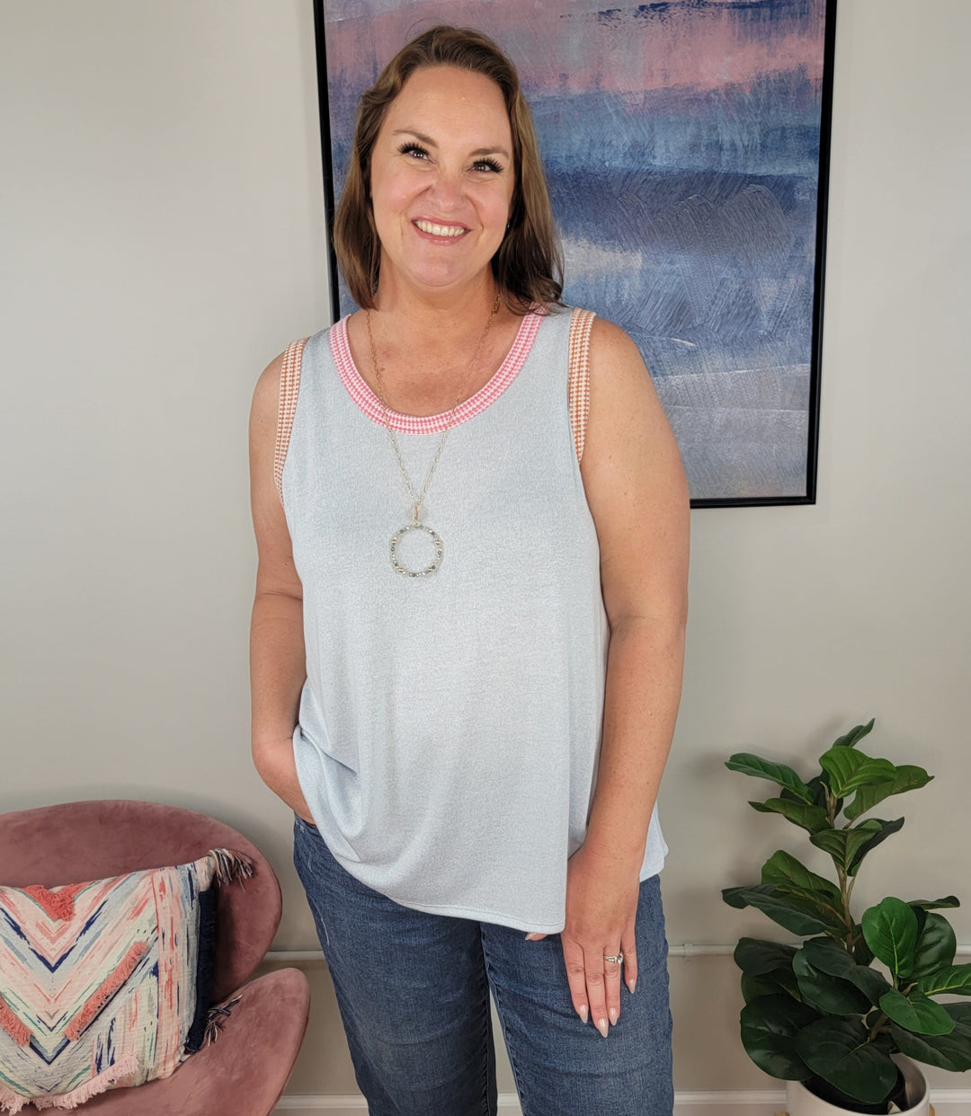 A smiling woman with shoulder-length brown hair models the Rousseau Vida Tank in light blue and jeans, standing indoors by a pink chair, patterned pillow, green plant, and a modern abstract painting on the wall.