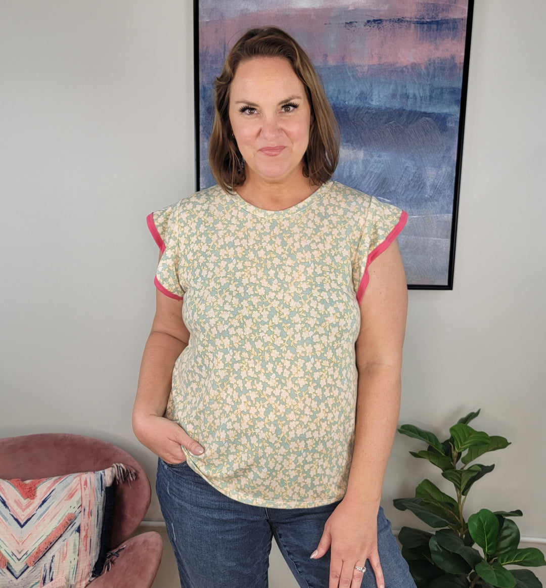 A woman with shoulder-length brown hair stands indoors wearing the Rousseau Murphy Floral Top, featuring mint green fabric, coral piping, and flutter sleeves, paired with blue jeans. She poses near a pink chair, a patterned pillow, plant, and framed art.
