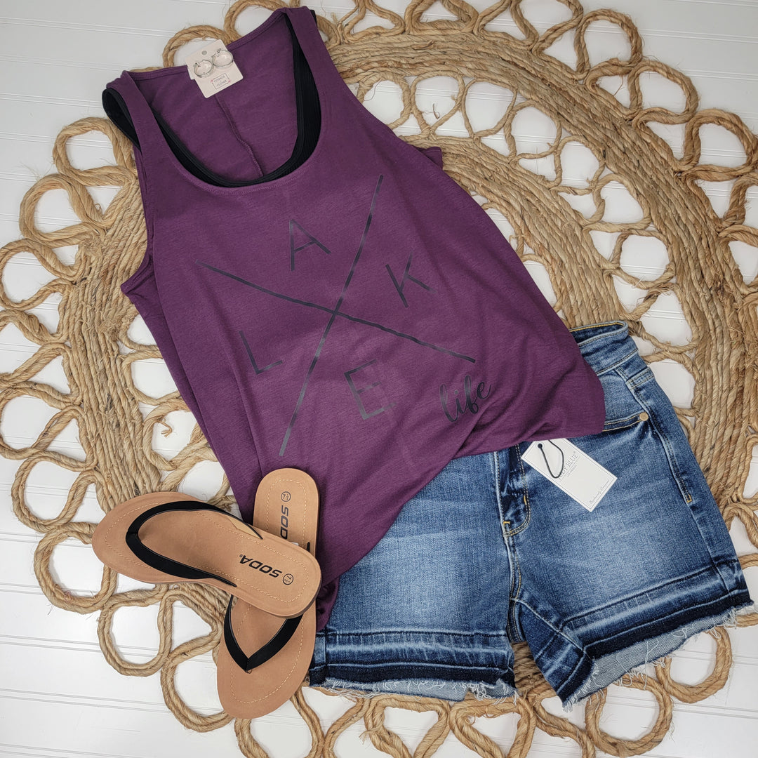 A Zenana Lake Life Graphic Tank in eggplant purple, denim shorts with a tag, and tan SODA flip-flops are displayed on a woven circular mat atop a white background.
