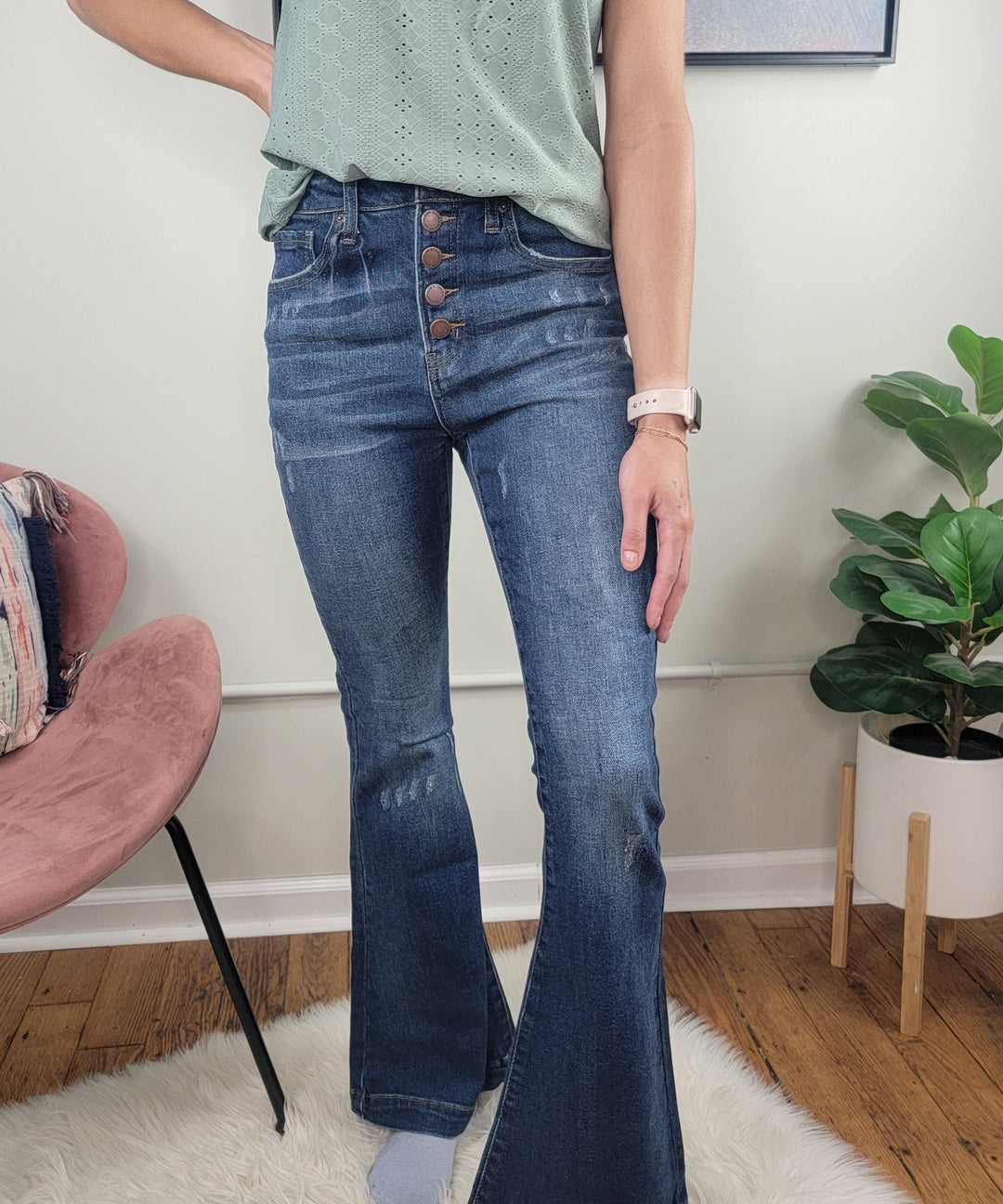 A woman’s lower half is shown as she stands indoors on a white rug beside a pink chair and potted plant, wearing Risen’s Coralie Vintage Wash Flares and a light green sleeveless top.