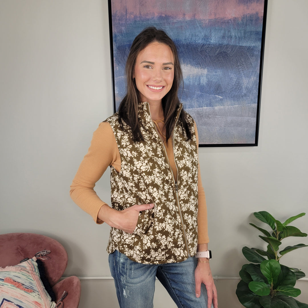 A smiling woman with long brown hair wears a tan long-sleeve shirt and the Staccato Chelsea Floral Vest, standing in a modern living room with a pink chair, pillow, green plant, and abstract painting on the wall.