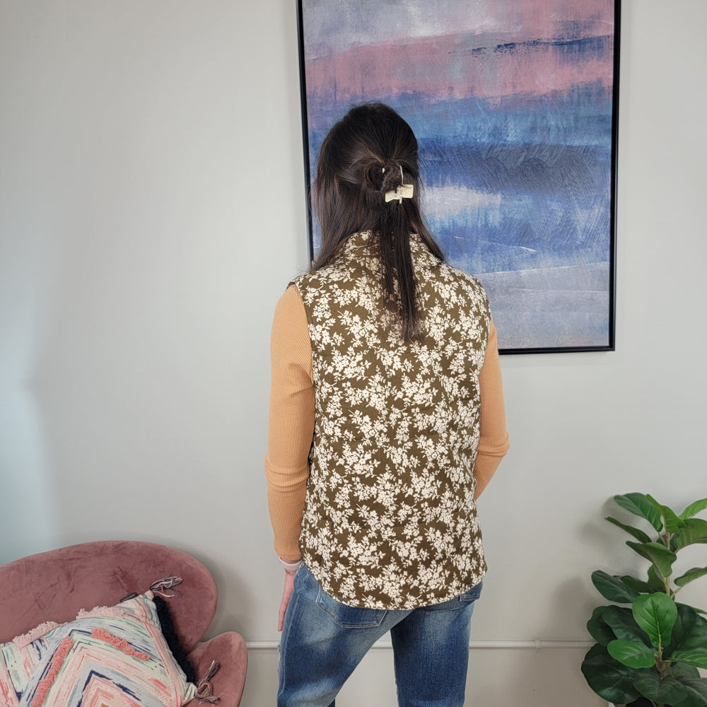 Wearing the Staccato Chelsea Floral Vest over a tan long-sleeve and blue jeans, a woman with long dark hair stands by an abstract painting. A pink chair and green plant nearby complete the cozy fall style.
