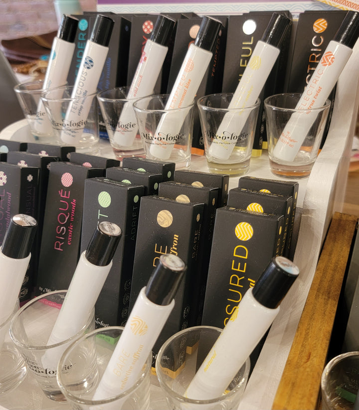 A display of Mixologie Blendable Perfume samples in glass vials stands in shot glasses, with Mixologie's colorful, alcohol-free boxes labeled with scent names arranged behind them.