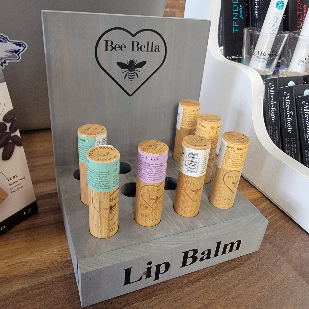 A wooden display stand labeled Bella Bee holds several bamboo tube Bella Bee Lip Balms made with Wisconsin beeswax and colored labels. The stand is topped by a heart-shaped Bella Bee logo featuring a bee icon.