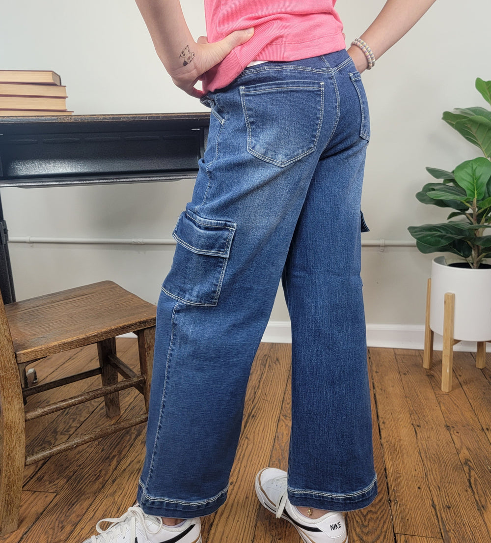A girl wears Cutie Patootie’s Bex Wide Leg Jeans, styled with a pink shirt and white sneakers. She stands indoors near a wooden chair, black desk, and potted plant; only the lower half of her body is visible.