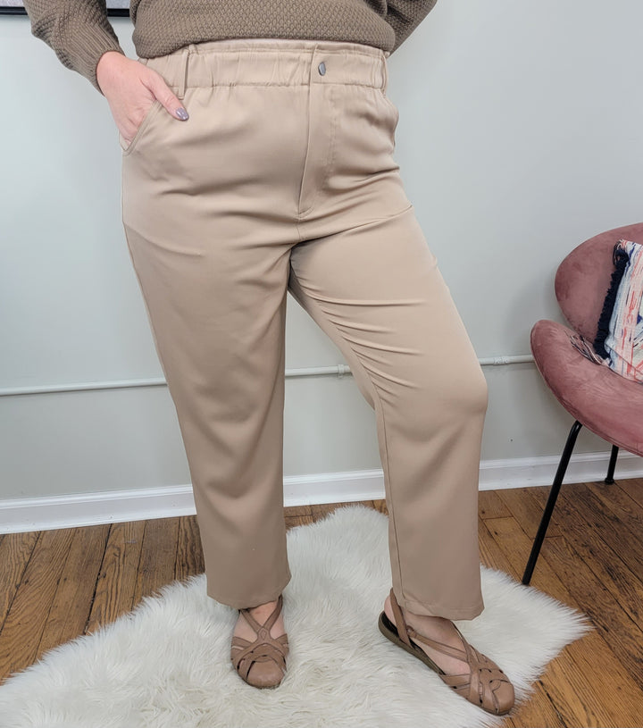 A person stands on a white rug wearing the ee:some Phoebe Dress Pants in beige with a brown sweater and tan sandals. One hand is in a pocket. A pink chair and wooden floor are visible in the background.