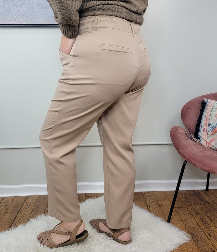 A person stands on a white rug wearing the ee:some Phoebe Dress Pants in light beige polyester with a high-waisted paper bag waistband, paired with a brown sweater and sandals. Their back is turned; a pink chair and white wall are behind them.