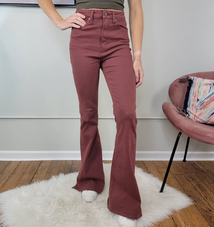 A person stands on a white rug wearing JUST USA Victoria Brick Flares in a maroon shade. Their upper body is cropped out; to the right is a pink chair with a colorful pillow. White shoes complete the look.