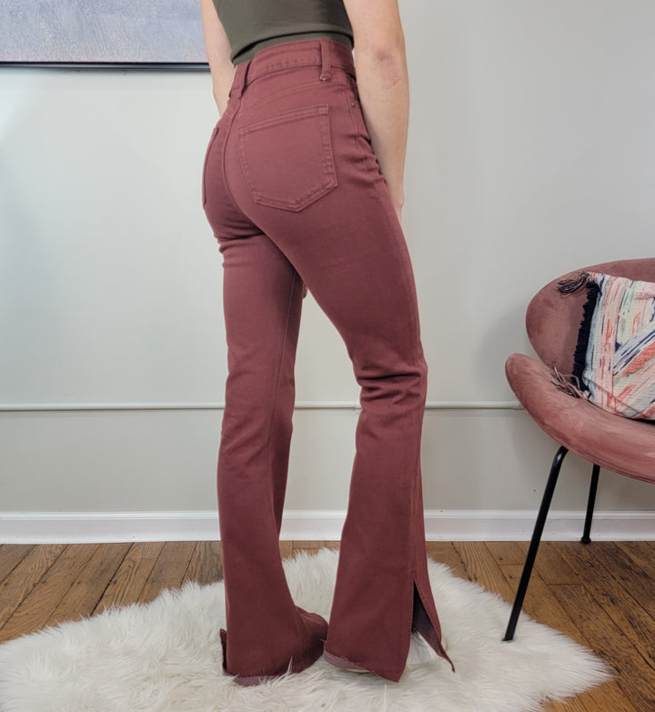 A person stands on a white rug wearing JUST USA’s Victoria Brick Flares—high-waisted, brick-red jeans with bottom slits. A pink chair with a patterned cushion and wood flooring are in the background.