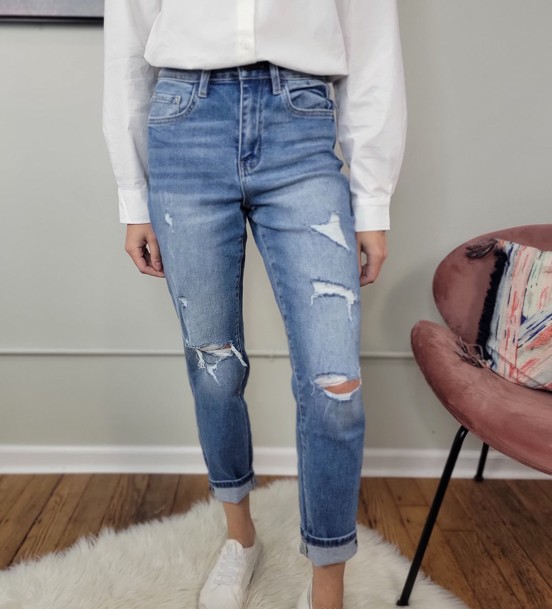 A person wearing a white long-sleeve shirt, Vervet Izzie Mom Jeans, and white sneakers stands on a white rug near a pink chair in a room with hardwood floors.