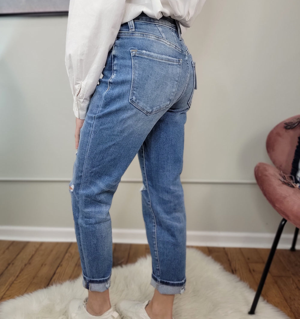 Wearing Vervet Izzie Mom Jeans in blue, a white long-sleeve shirt, and white sneakers, a person stands on a white rug with wooden floors and a pink chair nearby, turned to the side to show the high-rise jeans’ back.