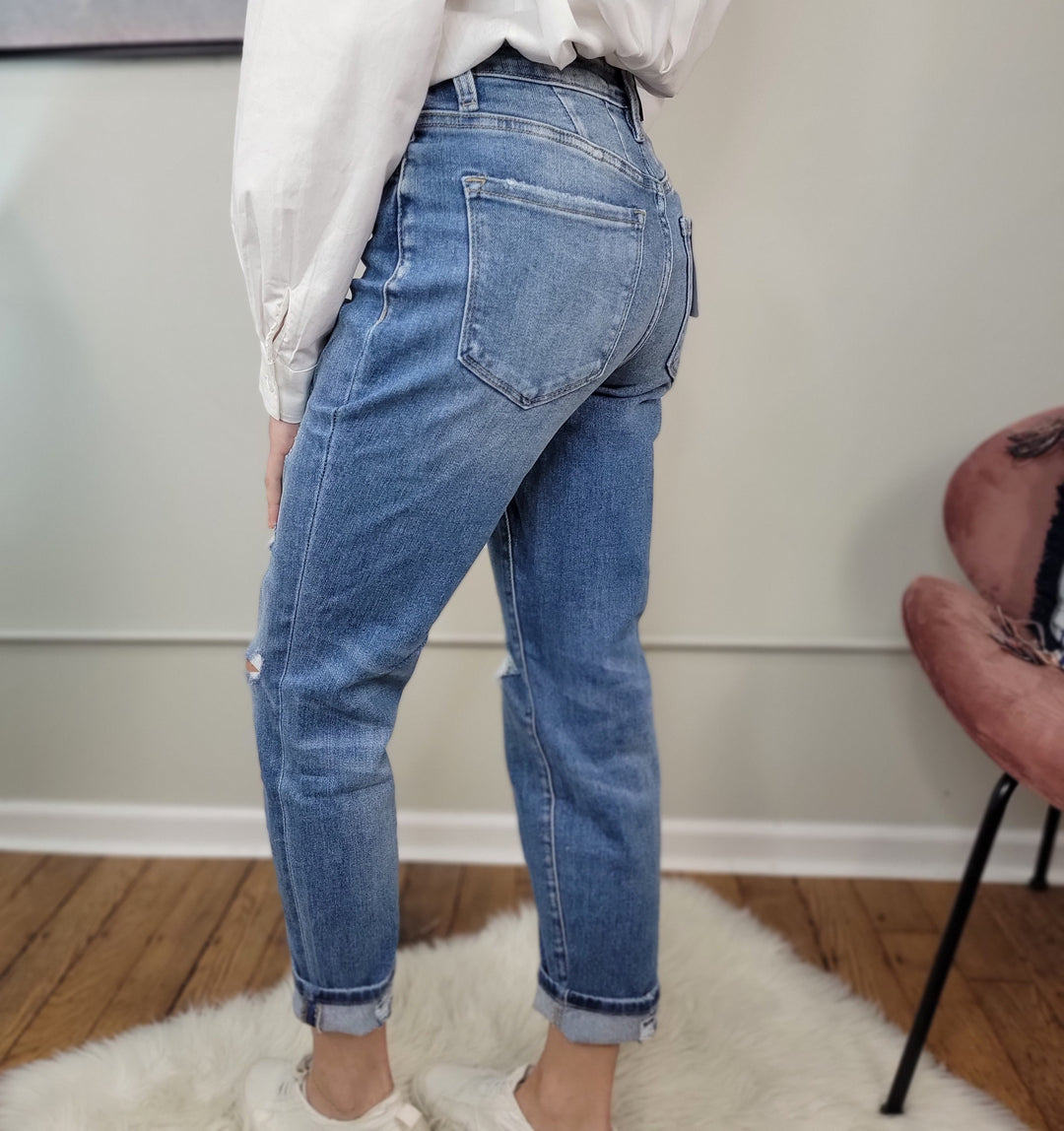 Wearing Vervet Izzie Mom Jeans in blue, a white long-sleeve shirt, and white sneakers, a person stands on a white rug with wooden floors and a pink chair nearby, turned to the side to show the high-rise jeans’ back.