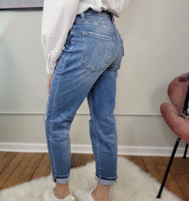 Wearing Vervet Izzie Mom Jeans in blue, a white long-sleeve shirt, and white sneakers, a person stands on a white rug with wooden floors and a pink chair nearby, turned to the side to show the high-rise jeans’ back.
