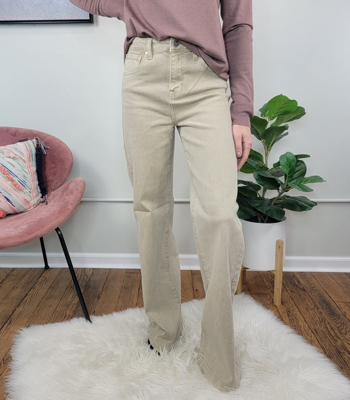 A person wears Risen's Lacie Wide Leg Jeans in light beige with a mauve long-sleeve top, standing on a white fuzzy rug beside a pink chair and potted plant, set against wooden floors and a light gray wall.