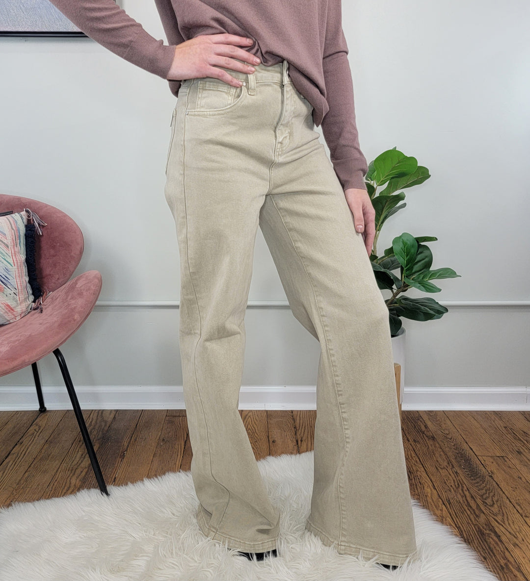 A person stands indoors on a white rug, wearing Risen's Lacie Wide Leg Jeans with tummy control and a mauve long-sleeve top, one hand on their hip. A pink chair and green plant are in the background.