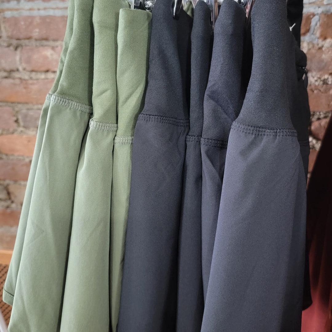Several pairs of Zenana Adrianne Shorts in olive green and black hang on hangers against an exposed brick wall, highlighting their functional high-waisted design with a convenient zipper back pocket.