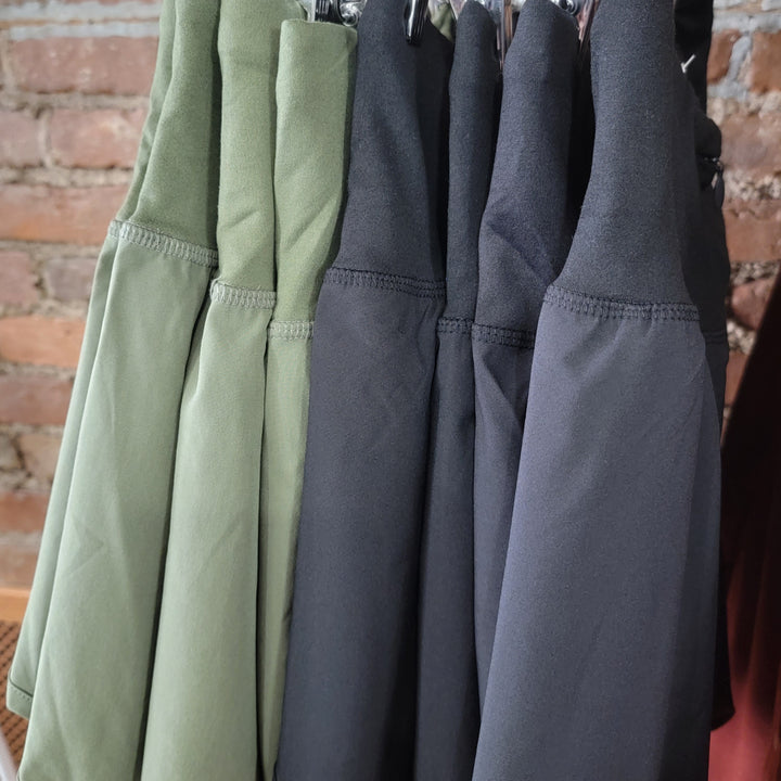 Several pairs of Zenana Adrianne Shorts in olive green and black hang on hangers against an exposed brick wall, highlighting their functional high-waisted design with a convenient zipper back pocket.