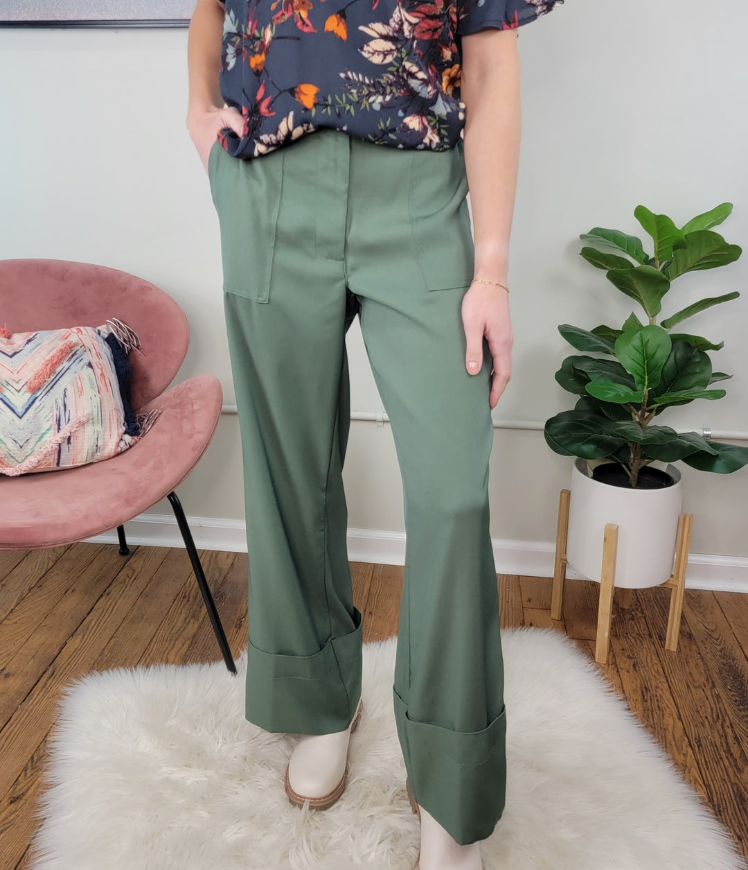 Indoors, a person models HYFVE, Inc's Erin Dress Pants in olive green with a dark floral top and cream shoes on a white rug. A pink chair, patterned pillow, and potted plant complete this stylish workspace look.