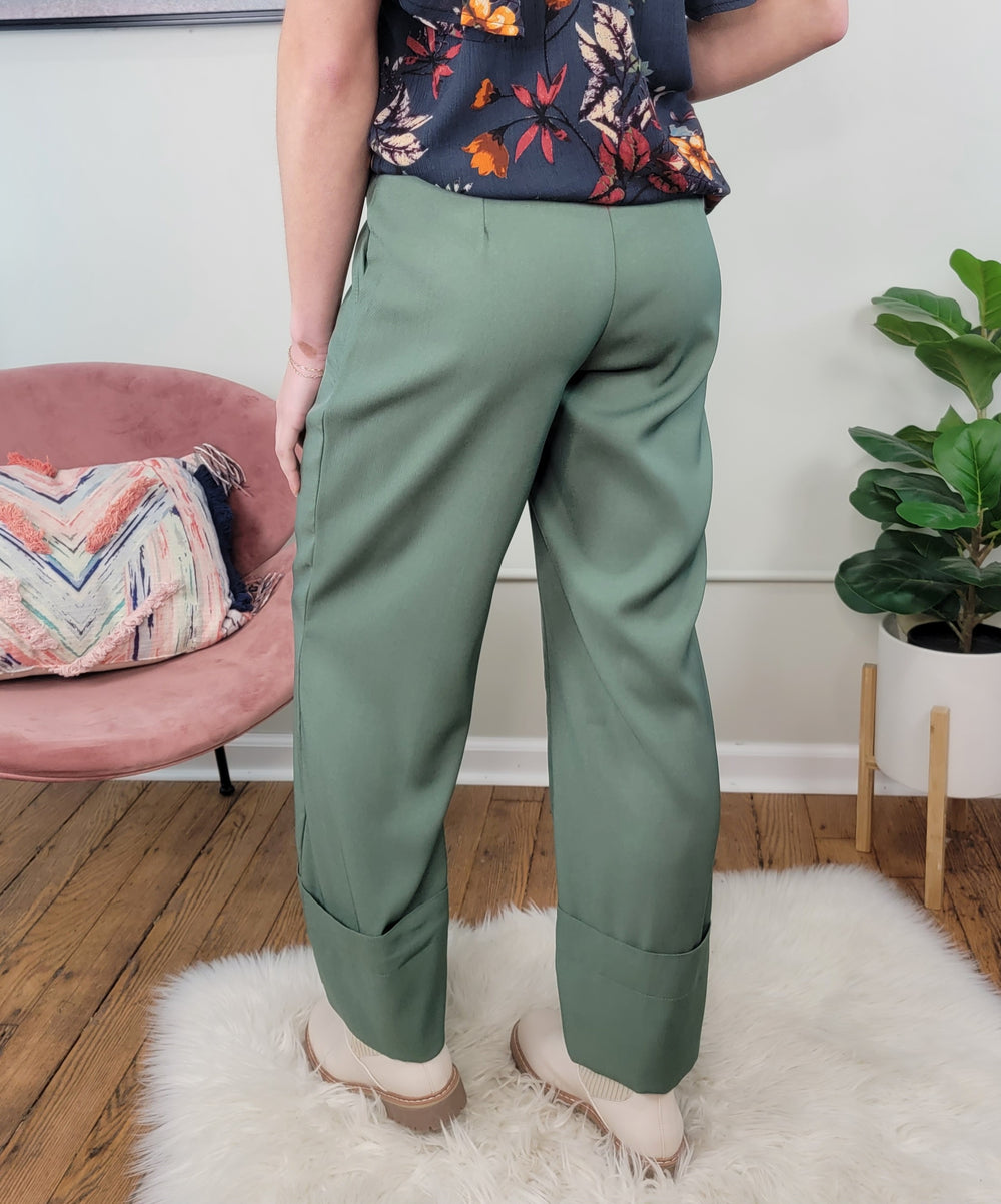 A person stands on a white rug in HYFVE, Inc’s Erin Dress Pants, paired with a dark floral top and beige shoes. A pink chair with a patterned pillow and a potted plant complete the stylish work wardrobe scene.