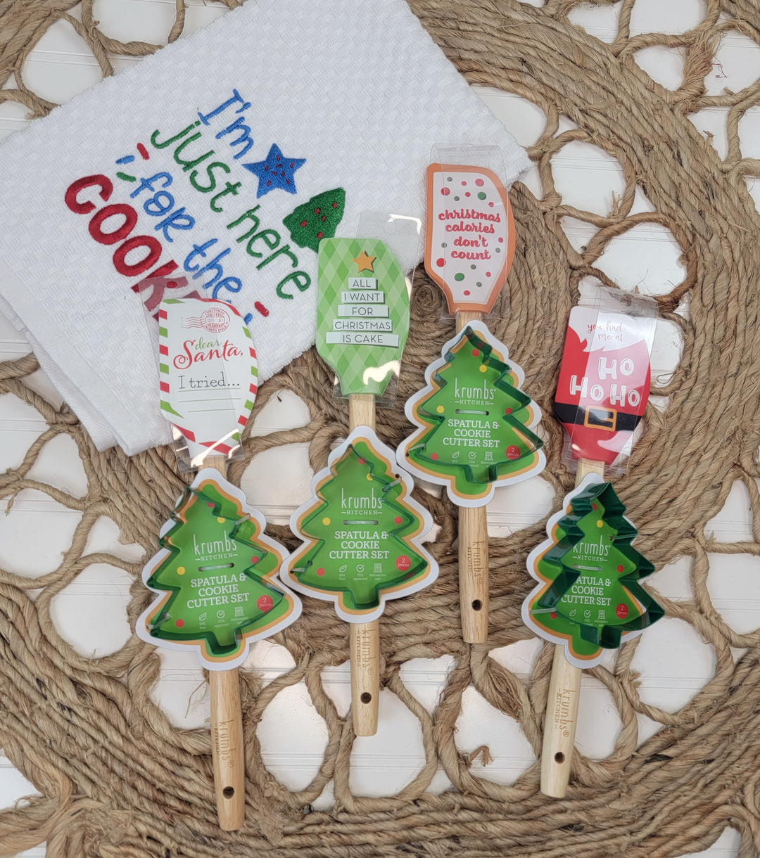 Five DM Merchandising Christmas Spatulas with Cookie Cutters, including three tree-shaped ones, are displayed on a woven mat below an embroidered tea towel that says "I’m just here for the cookies," creating a festive holiday scene.