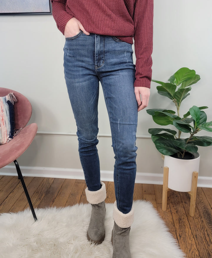 A person in a red ribbed sweater, Judy Blue Imogen Tummy Control Skinny Jeans, and gray shearling boots stands on a white rug beside a pink chair and potted plant in a cozy indoor setting.