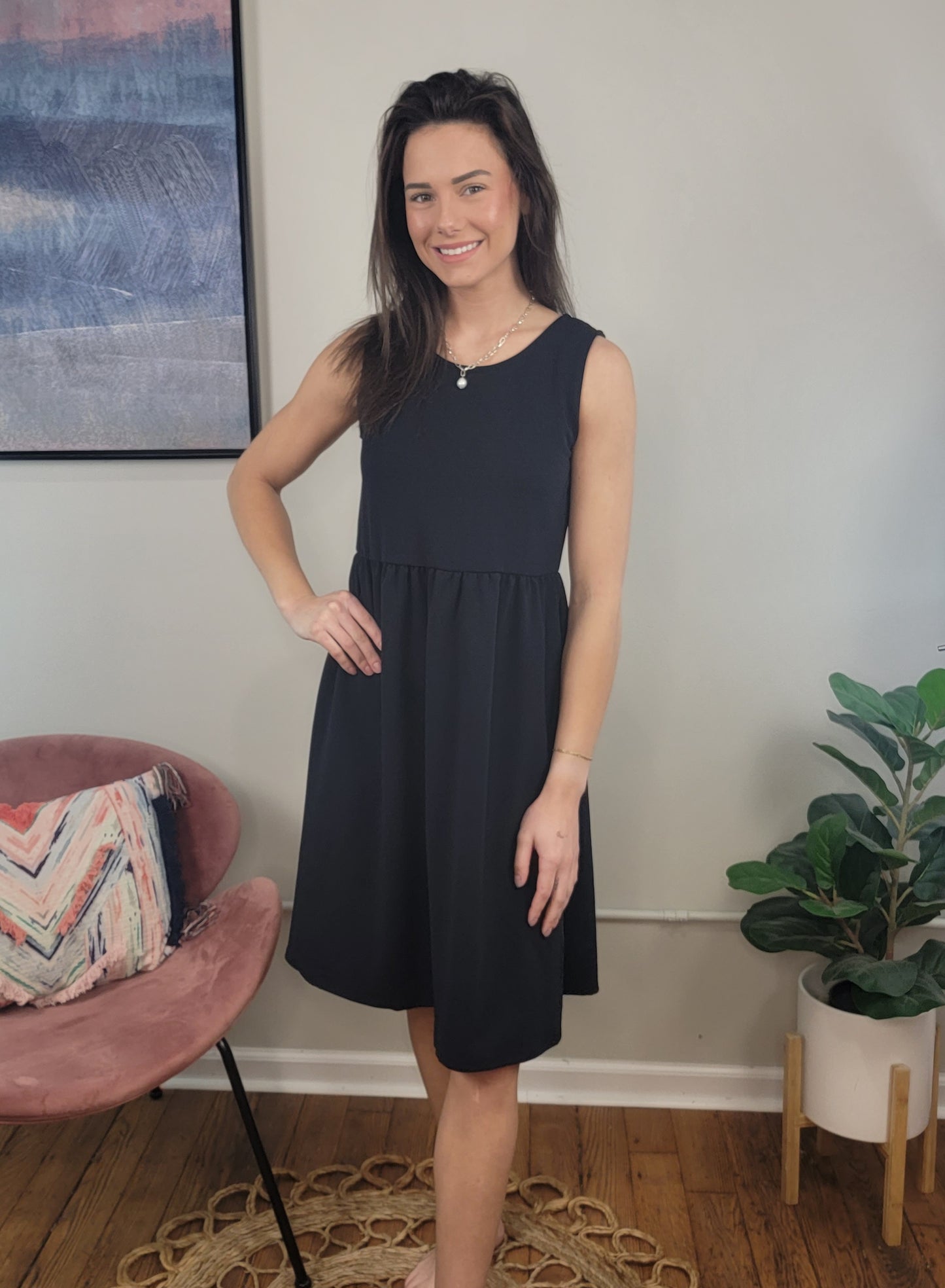 A woman with long brown hair smiles indoors, wearing the sleeveless Harmony Dress by Chris & Carol Apparel. She stands beside a pink chair with a patterned pillow and a green potted plant on the wooden floor.