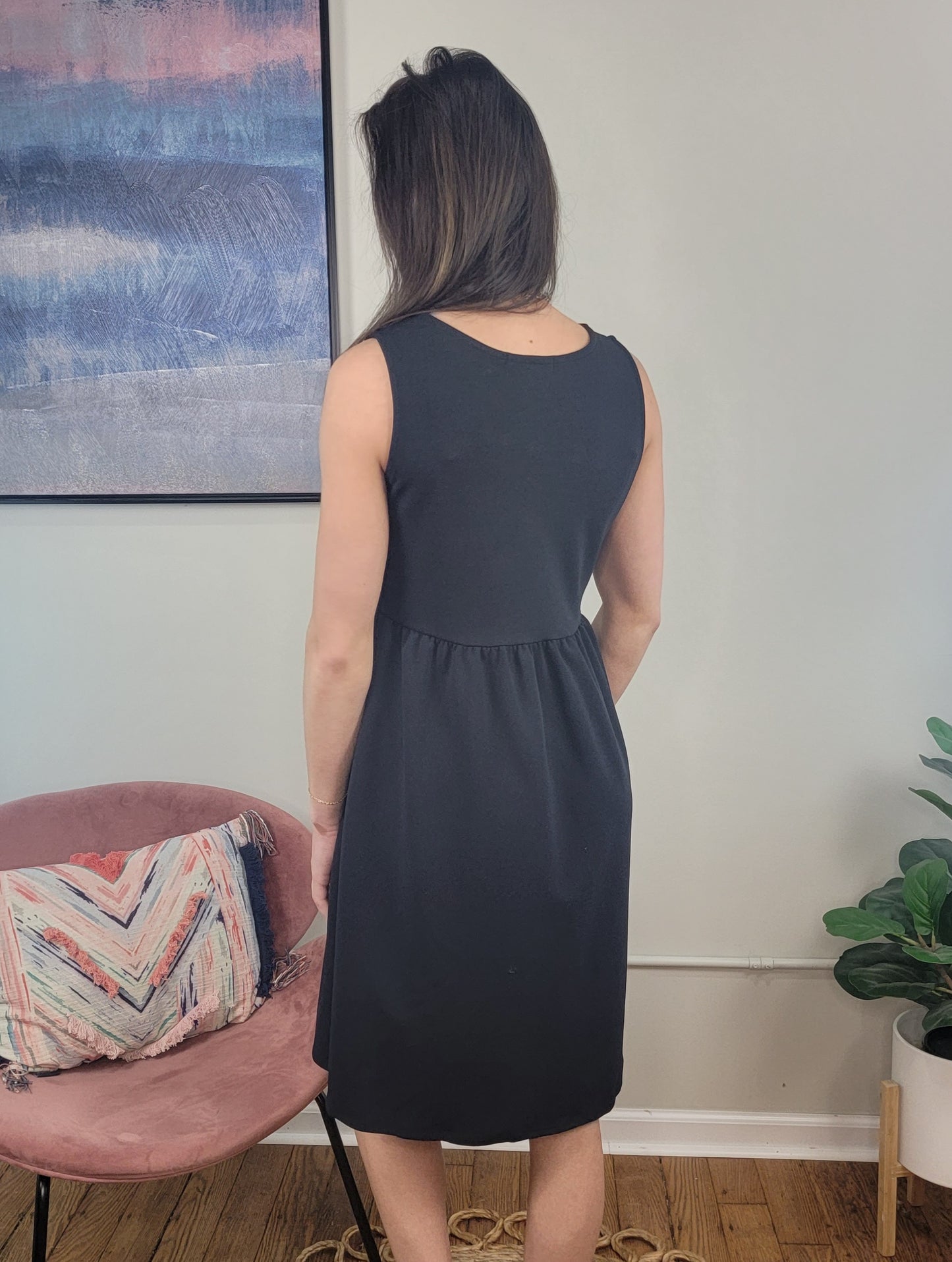 A woman with long brown hair stands indoors, facing away while wearing the Harmony Dress by Chris & Carol Apparel. She is near a pink chair with a patterned cushion, a green plant, and abstract art on the wall.