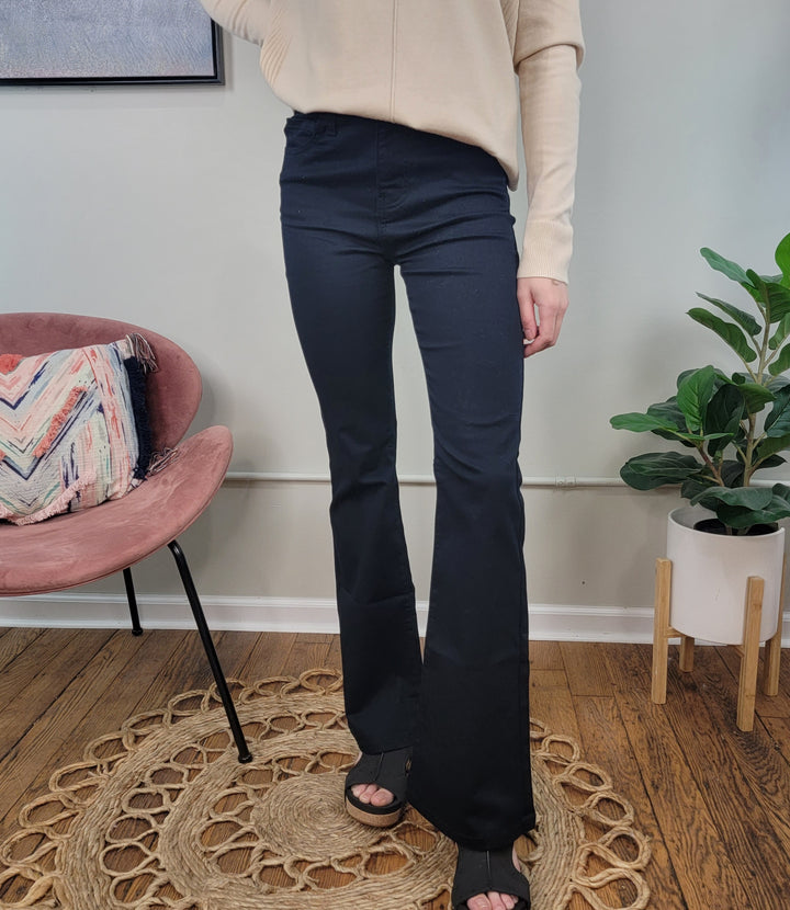Indoors, a person wears Zenana Amanda Bootcut Jeans in black with a beige long-sleeve top and black sandals. A pink chair with a patterned cushion and a green potted plant sit on a wooden floor with a decorative rug nearby.